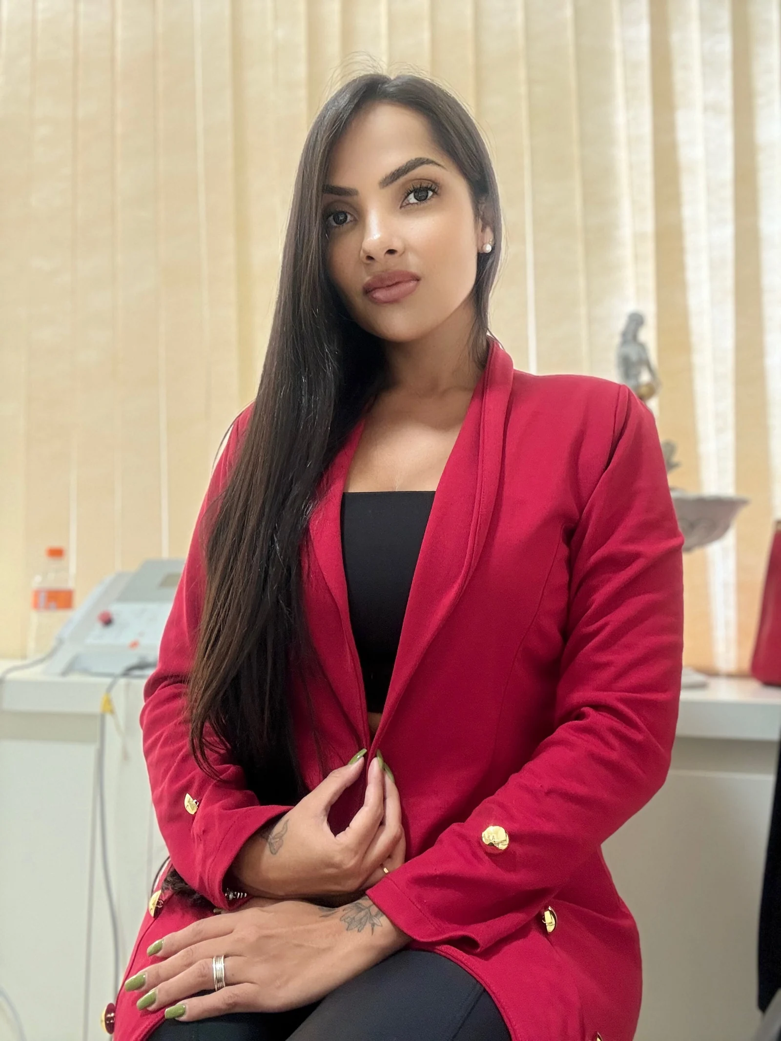 Dra. Fernanda Alves sentada na sala de atendimento, de blazer vermelho, diante da persiana creme com a luz do dia filtrada
