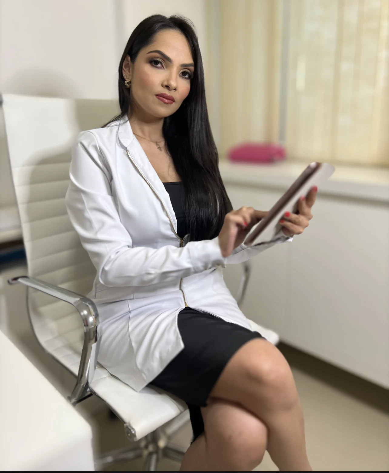 Dra. Fernanda Alves de jaleco branco, sentada com um tablet na sala de atendimento