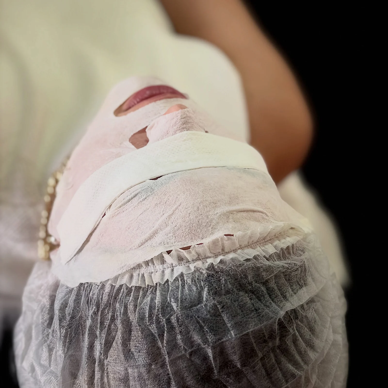 Paciente deitada com máscara facial e touca descartável durante um cuidado de pele na clínica